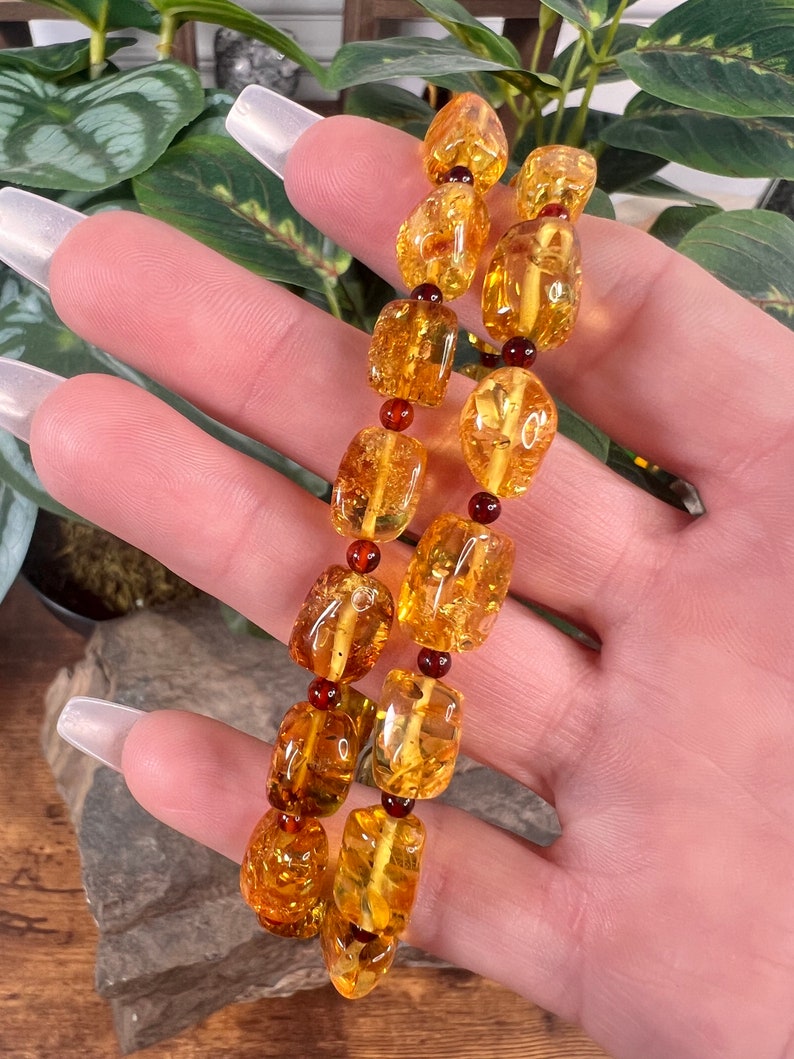 May include: A pair of amber bracelets with a warm, golden hue. The bracelets are made of irregularly shaped amber beads, interspersed with small, dark red beads. The bracelets are displayed against a backdrop of green foliage.