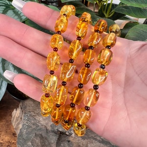 May include: Four strands of amber-colored beads with a polished, irregular shape. The beads are interspersed with small, dark red beads. The jewelry is displayed against a backdrop of green foliage and a natural stone.