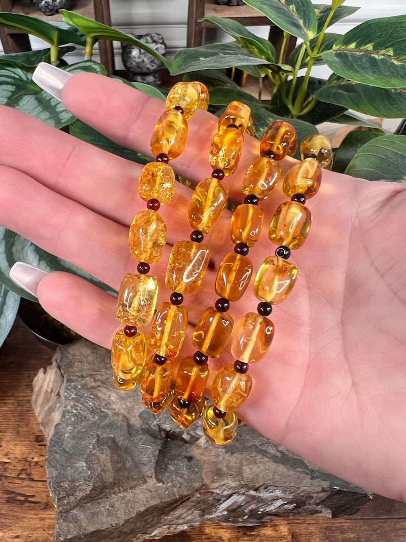 May include: Four strands of amber-colored beads with a polished, irregular shape. The beads are interspersed with small, dark red beads. The jewelry is displayed against a backdrop of green foliage and a natural stone.