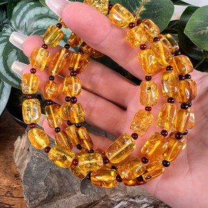 May include: A necklace featuring amber-colored, rectangular beads strung together with dark red and black accents. The beads have a polished, translucent appearance, and the necklace is displayed against a natural background.