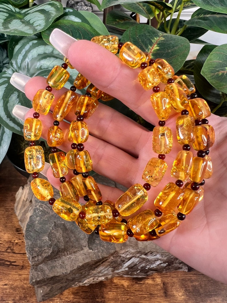 May include: A necklace featuring amber-colored, rectangular beads strung together with dark red and black accents. The beads have a polished, translucent appearance, and the necklace is displayed against a natural background.