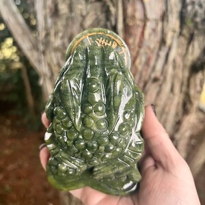 May include: A green jade frog sculpture with a coin on its head. The frog has detailed carvings and a smooth, polished surface. The coin has gold-colored accents and symbols. The sculpture is held in a hand against a blurred natural background.