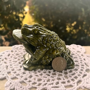 May include: A detailed, green frog sculpture with a coin in its mouth. The frog is positioned on a white, crocheted doily. A U.S. quarter is next to the frog for size comparison. The background is blurred.