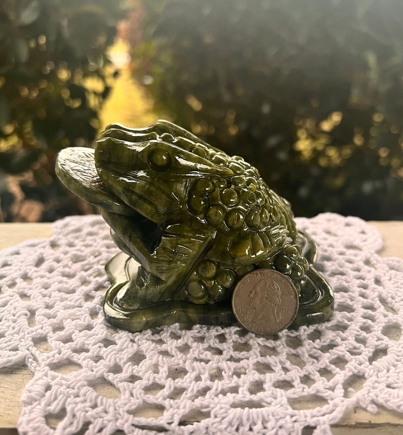 May include: A detailed, green frog sculpture with a coin in its mouth. The frog is positioned on a white, crocheted doily. A U.S. quarter is next to the frog for size comparison. The background is blurred.