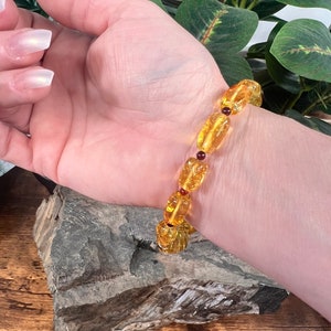 May include: A golden amber bracelet with oval-shaped beads and small red accent beads. The bracelet is worn on a wrist, showcasing the warm tones of the amber against the skin. The bracelet is a piece of jewelry.