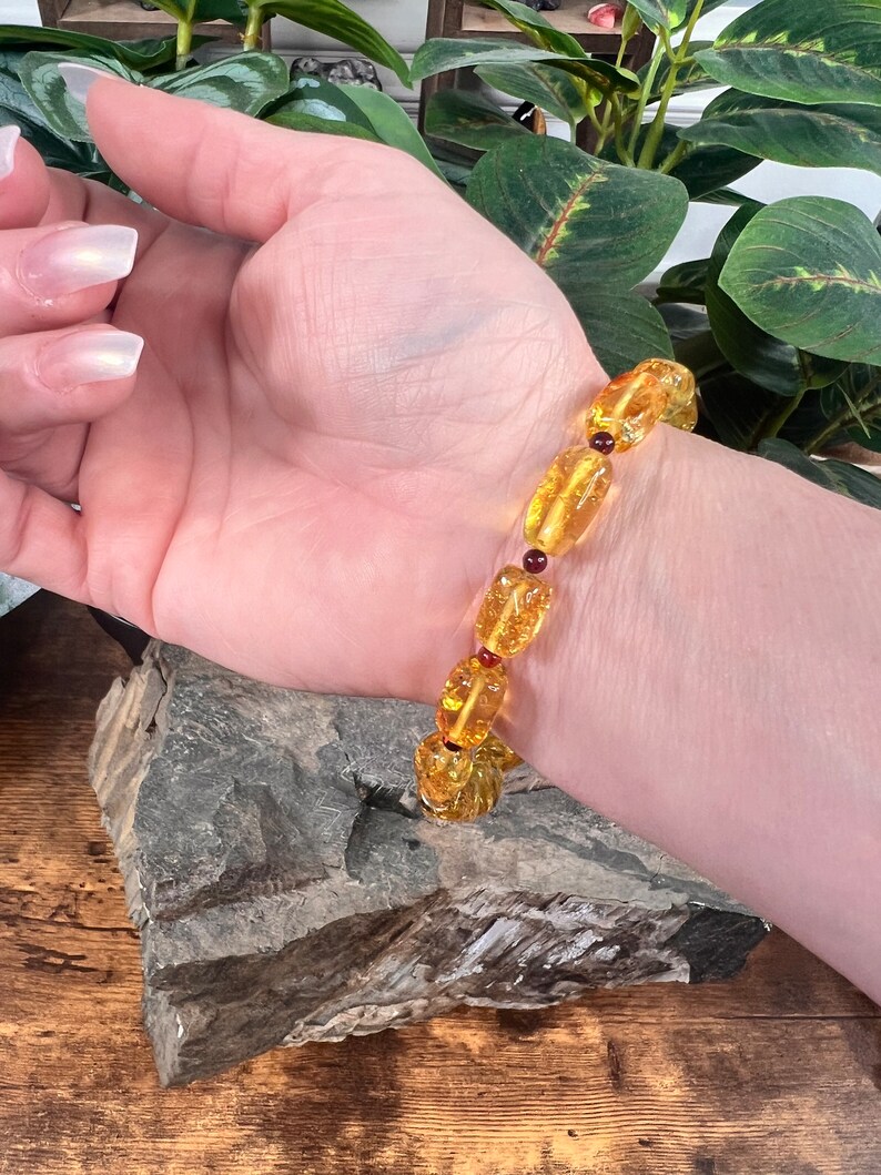 May include: A golden amber bracelet with oval-shaped beads and small red accent beads. The bracelet is worn on a wrist, showcasing the warm tones of the amber against the skin. The bracelet is a piece of jewelry.