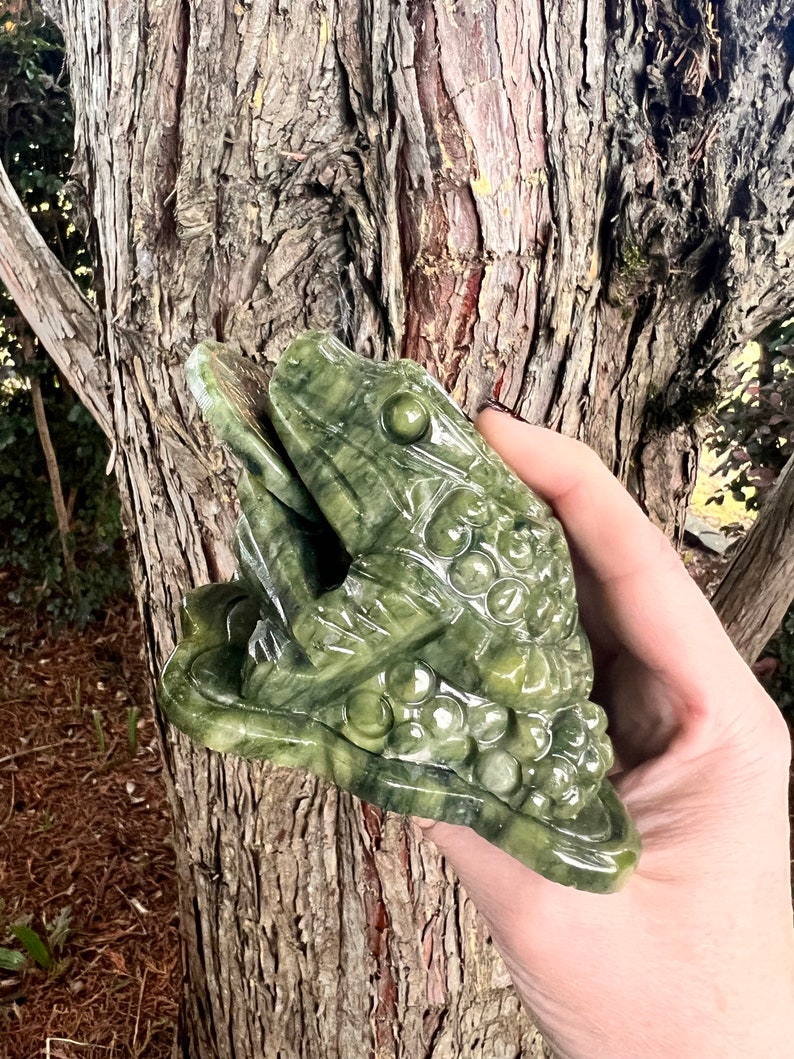May include: A green stone frog sculpture with detailed carvings, resting on a lily pad. The frog has textured skin and prominent eyes. The sculpture is held in a hand against a blurred tree bark background.