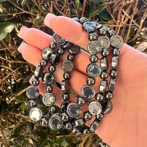 May include: A set of three dark gray beaded bracelets. Each bracelet features a mix of round and flat, disc-shaped beads. The bracelets are held in a hand, with a backdrop of green foliage.