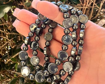 May include: A set of three dark gray beaded bracelets. Each bracelet features a mix of round and flat, disc-shaped beads. The bracelets are held in a hand, with a backdrop of green foliage.