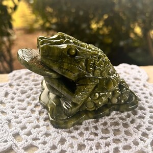 May include: A detailed, green jade frog sculpture with a coin in its mouth. The frog is carved with intricate details and sits on a decorative base. The sculpture is displayed on a white, lace doily.
