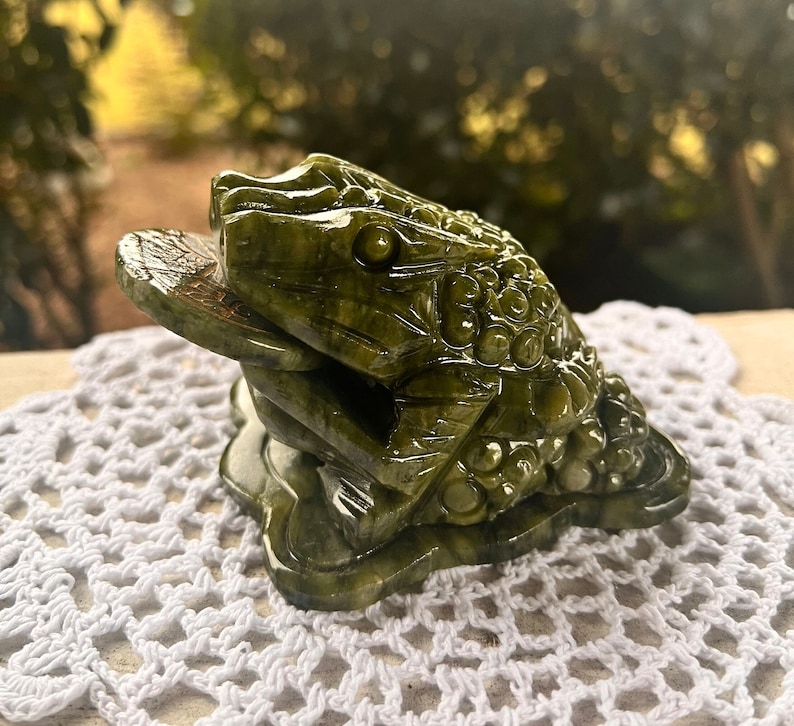 May include: A detailed, green jade frog sculpture with a coin in its mouth. The frog is carved with intricate details and sits on a decorative base. The sculpture is displayed on a white, lace doily.