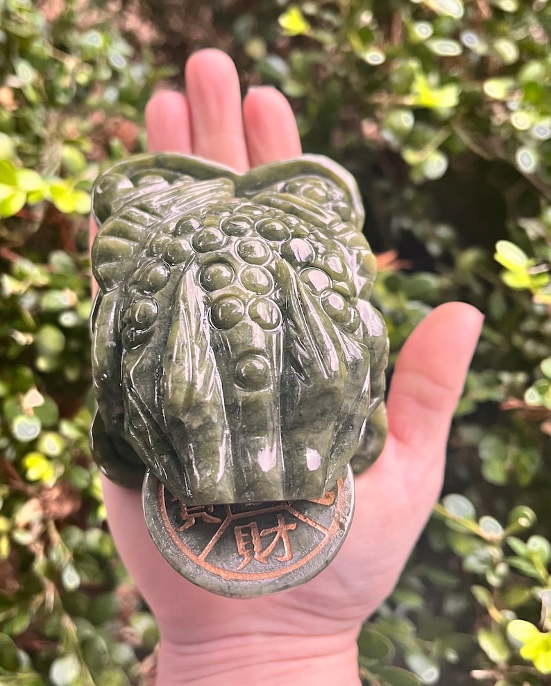 May include: A detailed, green jade frog sculpture holding a coin with Chinese characters. The frog is carved with intricate details, and the coin has a bronze-colored rim. The sculpture is held in a hand against a blurred green background.