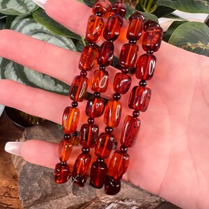 May include: A set of four amber-colored beaded bracelets. The beads are rectangular and oval-shaped, strung together with small black beads. The bracelets are displayed against a backdrop of green foliage and a wooden surface.