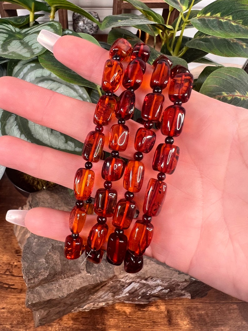 May include: A set of four amber-colored beaded bracelets. The beads are rectangular and oval-shaped, strung together with small black beads. The bracelets are displayed against a backdrop of green foliage and a wooden surface.