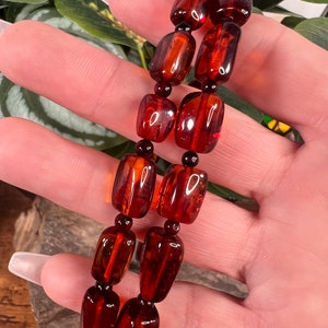 May include: A two-strand bracelet featuring rectangular amber beads with a rich, reddish-brown hue. The beads are separated by small, dark beads. The bracelet is held against a backdrop of green foliage.