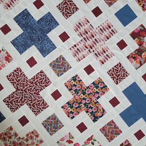 May include: A handmade quilt with a white background and a repeating pattern of colorful crosses. The crosses are filled with floral, geometric, and paisley designs in shades of blue, red, pink, and yellow. The quilt is a cozy and decorative home textile.