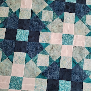 Peut inclure: Une courtepointe présentant un motif géométrique composé de pièces de tissu carrées et triangulaires. Le design utilise une palette de couleurs bleu marine foncé, sarcelle et blanc, avec divers imprimés batik incluant des motifs de feuilles et de fleurs.