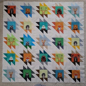May include: A white quilt featuring a repeating pattern of colorful squares with woodland animal illustrations. The quilt includes images of a moose, bear, deer, raccoon, eagle, fox, owl, and rabbit. The quilt is approximately 60 inches by 70 inches.