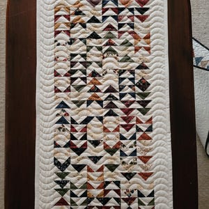 Peut inclure: Un chemin de table matelassé rectangulaire présentant un motif répétitif de petits triangles de différentes couleurs, notamment rouge, bleu, vert et crème. Le motif central est encadré par une bordure crème unie avec un matelassage ondulé.