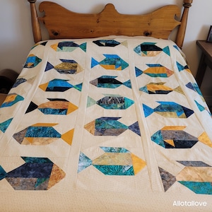 May include: A quilt featuring a repeating pattern of stylised fish in shades of blue, teal, gold, and black, set against a cream-coloured background. The fish are arranged in vertical columns separated by cream fabric strips. The text Allotallove is visible.
