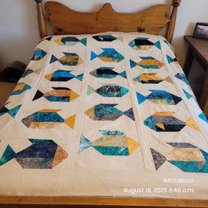 May include: A beige quilt with a repeating pattern of colourful fish. The fish are made of geometric shapes in shades of blue, teal, gold, and brown. The quilt is on a wooden bed frame. The text "Allotallove" and the date and time are visible.