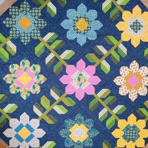 May include: A quilt featuring a dark blue denim-textured background with a repeating pattern of multi-coloured fabric flowers. Each flower has eight petals in various shades of yellow, pink, blue, and white, connected by green stems and leaves.