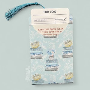 May include: A light blue TBR (To Be Read) log bookmark with a teal tassel. The bookmark features a repeating pattern of stacked books, crowns, and floral arrangements. Text on the bookmark includes "TBR LOG" and "KEEP THIS BOOK CLEAN".