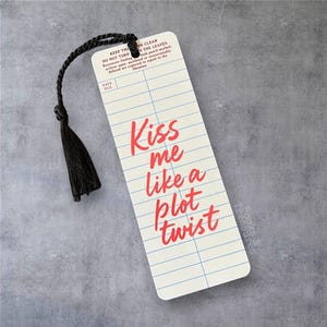 Peut inclure: Un marque-page rectangulaire avec un pompon noir. Le marque-page a un fond crème avec des lignes bleues et un texte rouge qui dit "Kiss me like a plot twist". Le haut du marque-page porte l'inscription "KEEP THE BOOK CLEAN".