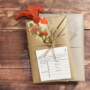 Könnte beinhalten: Zwei eingepackte Bücher mit einem dekorativen Etikett mit Pilzen und Herbstlaub. Die Bücher sind in braunes Papier gewickelt und mit Bindfaden verschnürt. Das Etikett listet die Genres des Buches als Mystery, Crime und Thriller auf.