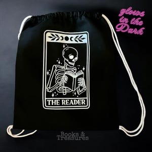 Può includere: Zaino nero con coulisse con una grafica bianca di uno scheletro che legge un libro, con la scritta "THE READER". La borsa ha coulisse bianche e il testo "glows in the Dark" e "Books & Treasures."