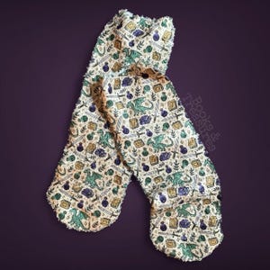 May include: Cream-colored oven mitts with a fantasy-themed pattern. The design includes green dragons, swords, potion bottles, and spell books. The mitts have a textured, fringed edge and are set against a dark purple background.