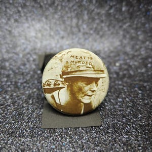 Puede incluir: Un botón redondo en tonos sepia con el texto "MEAT IS MURDER" sobre un retrato de una persona con sombrero. El botón está sobre un fondo gris oscuro.