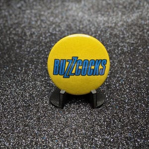 Pode incluir: Um crachá redondo amarelo com a palavra BUZZCOCKS impressa em letras maiúsculas azuis em negrito. O crachá é mantido na vertical por um pequeno suporte de plástico preto, exibindo o texto claramente contra o fundo amarelo brilhante.
