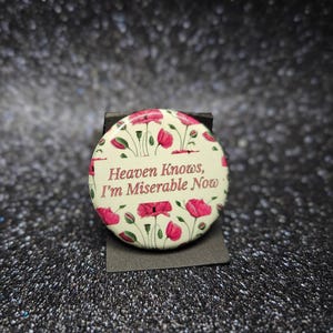 May include: A round button with a floral design featuring red poppies and green leaves. The button has the text "Heaven Knows, I'm Miserable Now" in a cream-colored banner.