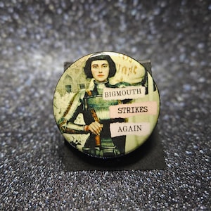 May include: A round button with a vintage illustration of a person in armor. The button has the text "BIGMOUTH STRIKES AGAIN" in a mix of fonts and colors. The background is a mix of beige and green.