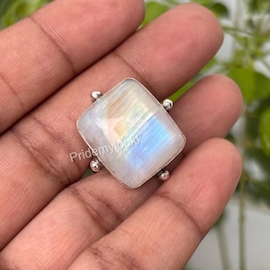 May include: A square-shaped ring with a large, iridescent moonstone. The stone displays a play of colors, including blue, yellow, and white. The ring has a silver band with small decorative accents. The text "Pridemylookk" is visible on the stone.
