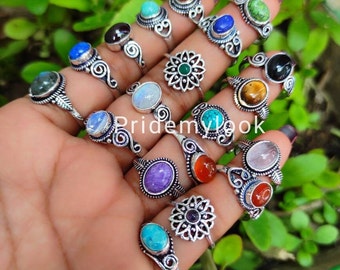 May include: A collection of silver rings with various gemstone settings, including blue, green, and red stones. The rings are displayed on a hand against a backdrop of green foliage. The rings have intricate designs.
