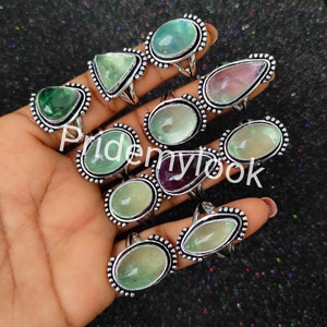 May include: A collection of 12 silver rings with green, purple, and clear gemstone settings. The rings are arranged on a hand, with the gemstones facing the camera. The rings are all oval-shaped and have a beaded edge.