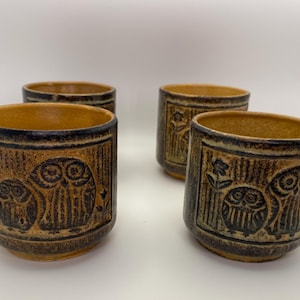 May include: Four ceramic owl-themed cups. The cups are cylindrical with a brown and tan color scheme. Each cup features an embossed owl design and a dark blue rim. The cups are approximately 3 inches tall.