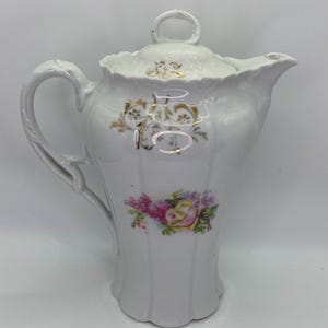 Può includere: Caffettiera in porcellana bianca con coperchio, manico e beccuccio. La caffettiera è decorata con accenti floreali dorati e un bouquet di fiori rosa, viola e gialli. Ha un bordo smerlato e un manico ad anello sul coperchio.