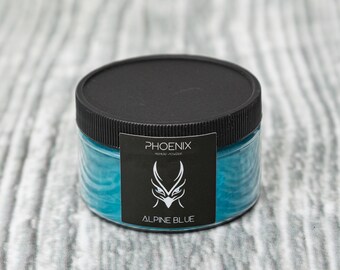 May include: A clear plastic jar with a black lid containing a blue pigment powder. The label reads "PHOENIX PIGMENT POWDER" and "ALPINE BLUE".