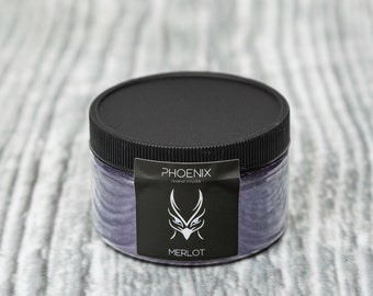 May include: A clear jar with a black lid containing a purple powder. The jar has a black label with the word "PHOENIX" and the word "MERLOT" printed on it. The label also features a white graphic of a stylized bird or mythical creature.
