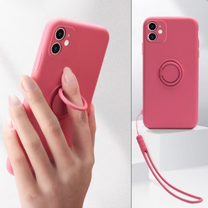 May include: A pink silicone phone case with a built-in ring holder and a detachable wrist strap. The case has a camera cutout and a raised lip around the screen to protect the phone from scratches.