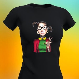 May include: Black t-shirt with a colorful illustration of a woman with pigtails, glasses, and tattoos. She is wearing a red and green outfit and has a peace sign on her hand.