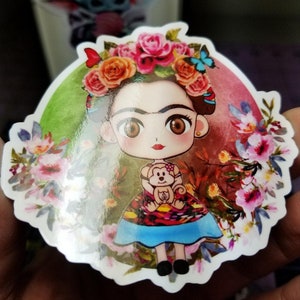 Peut inclure: Un autocollant color&eacute; repr&eacute;sentant une illustration de Frida Kahlo en dessin anim&eacute; avec un singe, entour&eacute;e de fleurs et d'un fond vert et rouge.