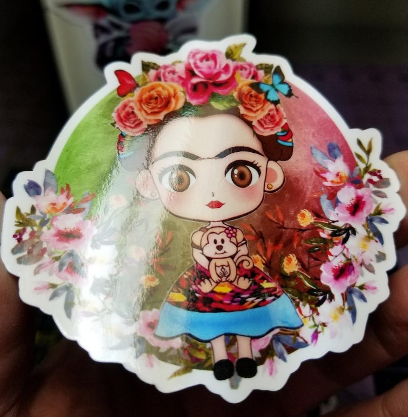 Peut inclure: Un autocollant color&eacute; repr&eacute;sentant une illustration de Frida Kahlo en dessin anim&eacute; avec un singe, entour&eacute;e de fleurs et d'un fond vert et rouge.