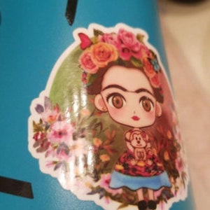 Peut inclure: Une gourde turquoise avec un autocollant repr&eacute;sentant une Frida Kahlo de dessin anim&eacute; portant une couronne de fleurs et tenant un singe. L'autocollant est entour&eacute; de fleurs et de papillons.