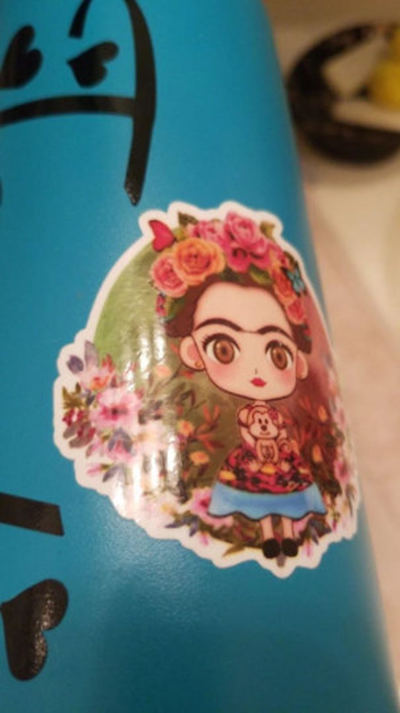 Peut inclure: Une gourde turquoise avec un autocollant repr&eacute;sentant une Frida Kahlo de dessin anim&eacute; portant une couronne de fleurs et tenant un singe. L'autocollant est entour&eacute; de fleurs et de papillons.