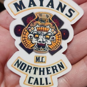 May include: A white sticker with the words "MAYANS" and "NORTHERN CALI." in black letters. The sticker features a detailed graphic of a Mayan-inspired skull with orange, yellow, and black accents, and the letters "M.C."