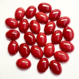 Può includere: Una collezione di gemme rosse lucide di forma ovale. Le gemme sono disposte in modo sparso su uno sfondo bianco. Ogni pietra ha una superficie liscia e lucida, che riflette la luce. L'impressione generale è quella di una collezione di pietre naturali vibranti.